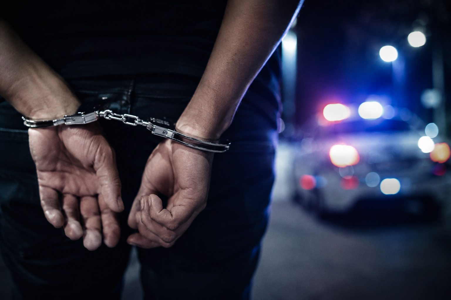 Mãos algemadas durante prisão em flagrante à noite, com luzes de viatura policial desfocadas ao fundo